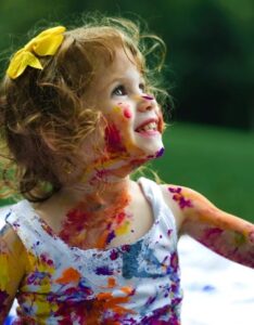 En ung flicka, ett barn runt 3 år, med ansiktsfärg och vattenfärg på armarna tittar upp och ler. Hon har mörkt hår som är lite lockigt.